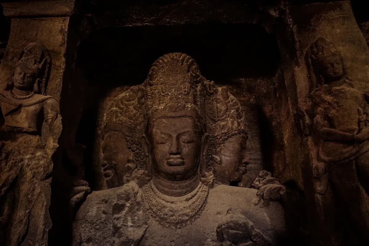 A large carved stone figure with three heads in a dark cave at Elephanta, flanked by carved standing figures