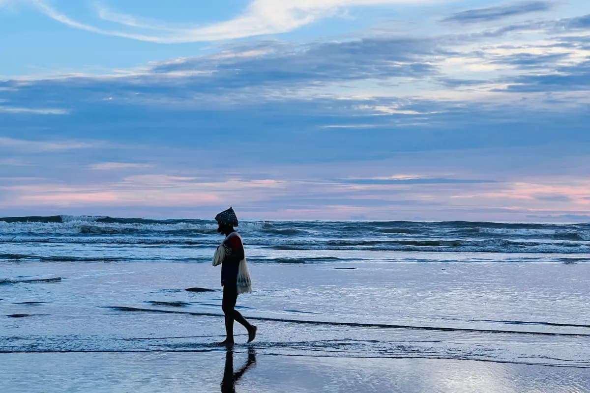 A man carrying a fishing net walks along the wet sand at dawn, with waves breaking behind
