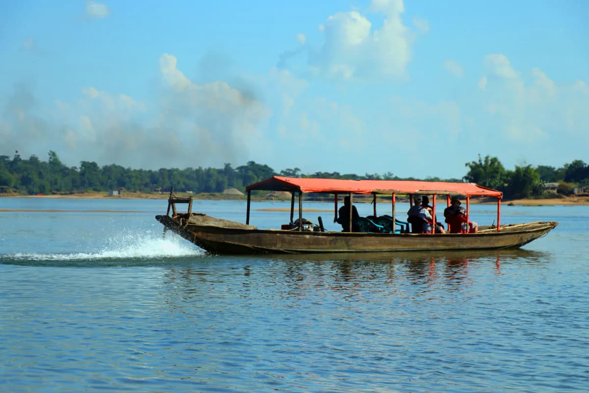 A wooden passenger boat with a red canopy crossing a wide river, with trees on the far bank