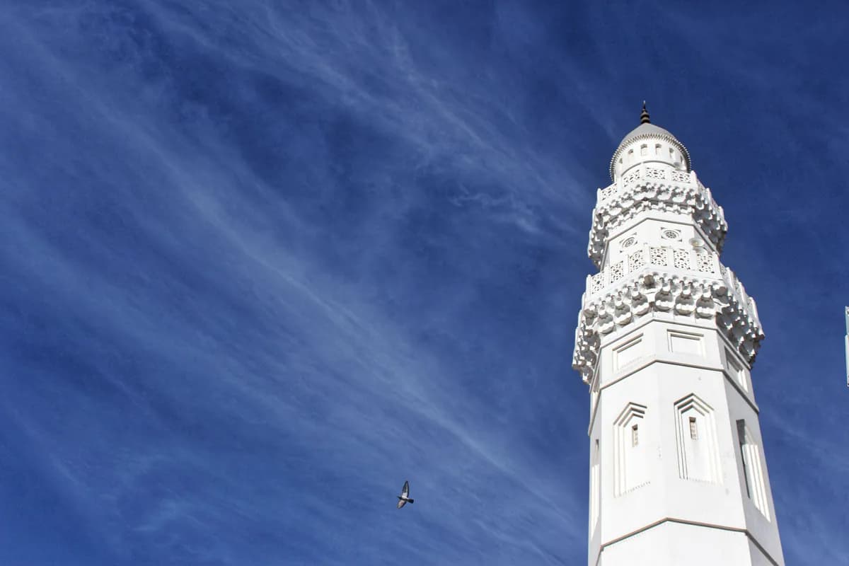 A white minaret of Quba Mosque against a blue sky with thin clouds and a pigeon in flight