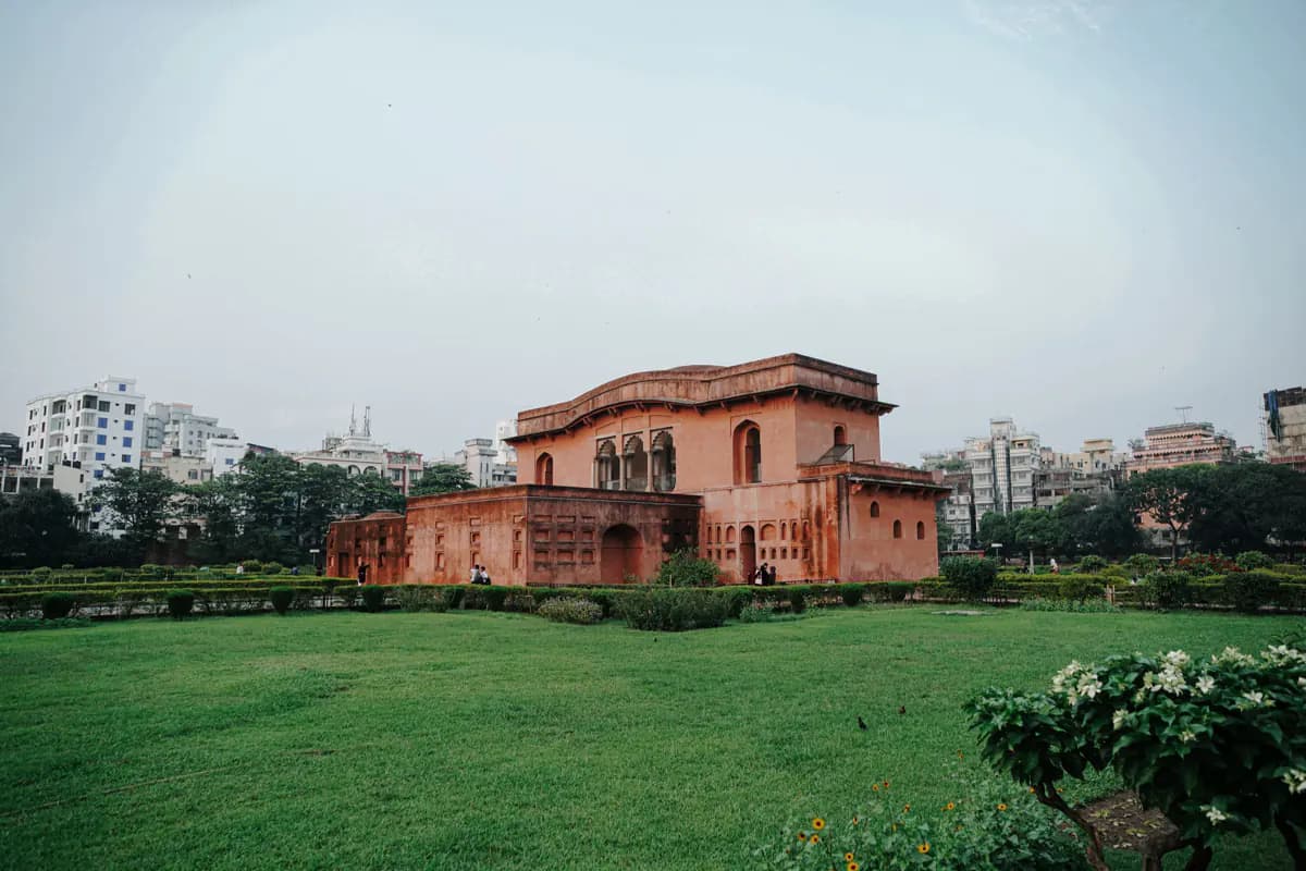 A red Mughal building with arched windows at Lalbagh Fort, behind lawns and hedges, with apartment blocks beyond